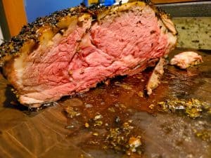A close-up of a juicy, medium-rare roast beef with a herb crust, sliced open to reveal a pink interior, resting on a cutting board with juices and seasonings.