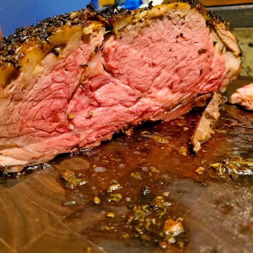 A close-up of a juicy, medium-rare roast beef with a herb crust, sliced open to reveal a pink interior, resting on a cutting board with juices and seasonings.