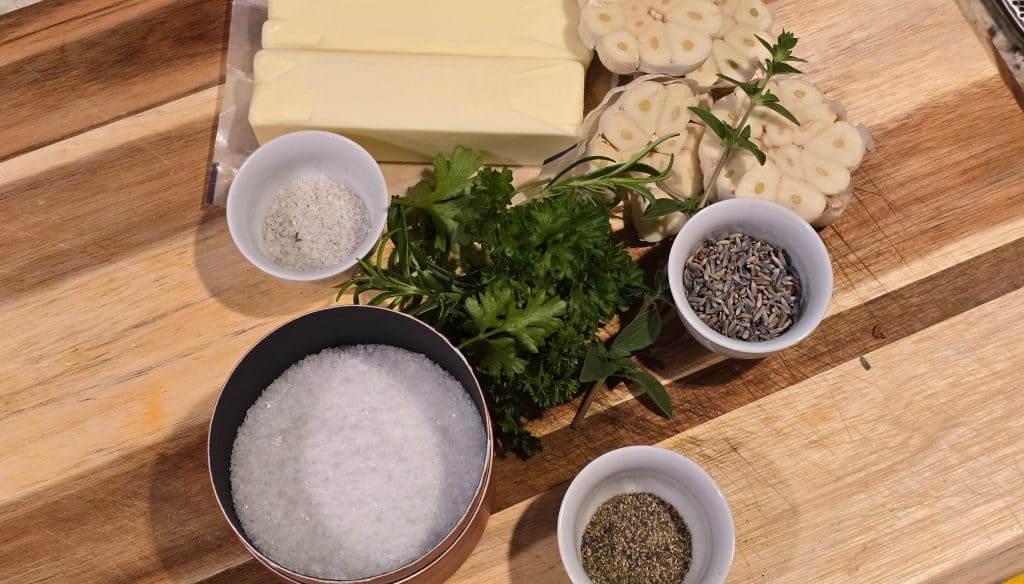 Sticks of butter, halved garlic bulbs, small bowls of salt, dried herbs, and pepper are arranged on a wooden cutting board with fresh parsley and tarragon in the center.