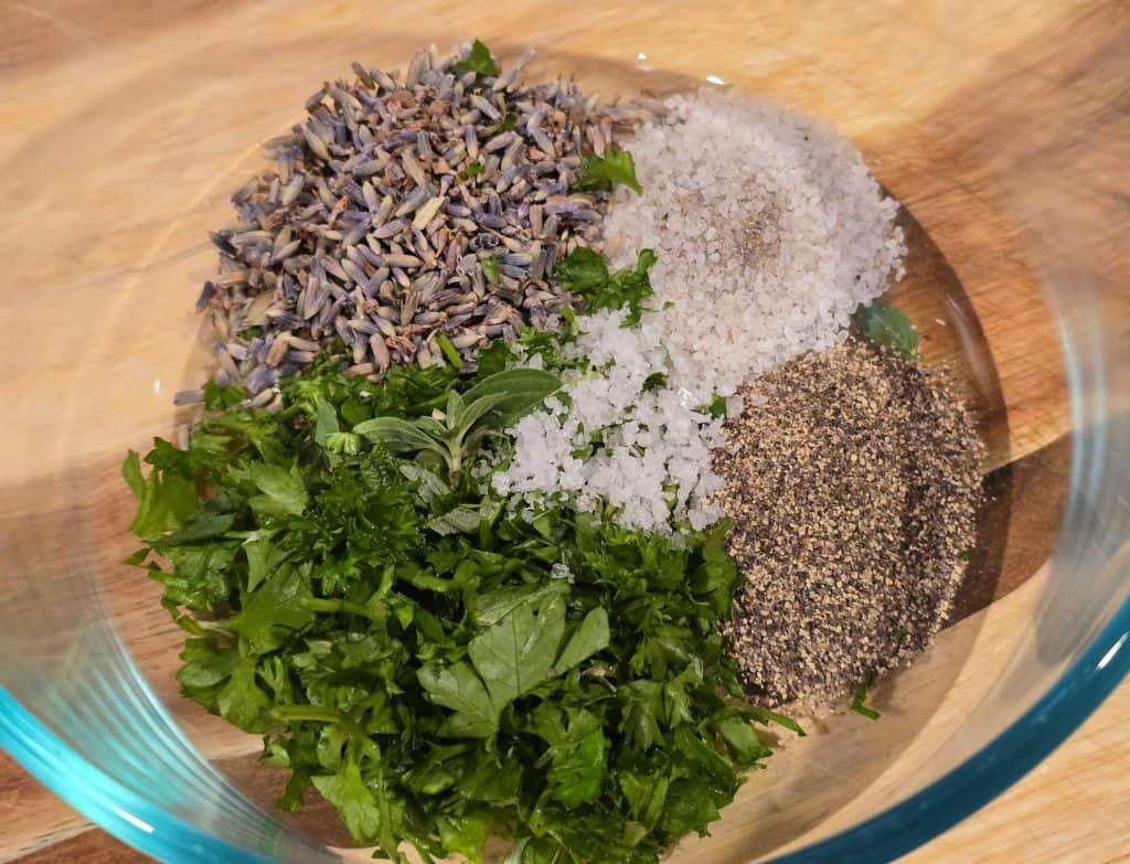 A glass bowl containing piles of dried lavender, coarse salt, chopped fresh parsley, and ground black pepper on a wooden surface.