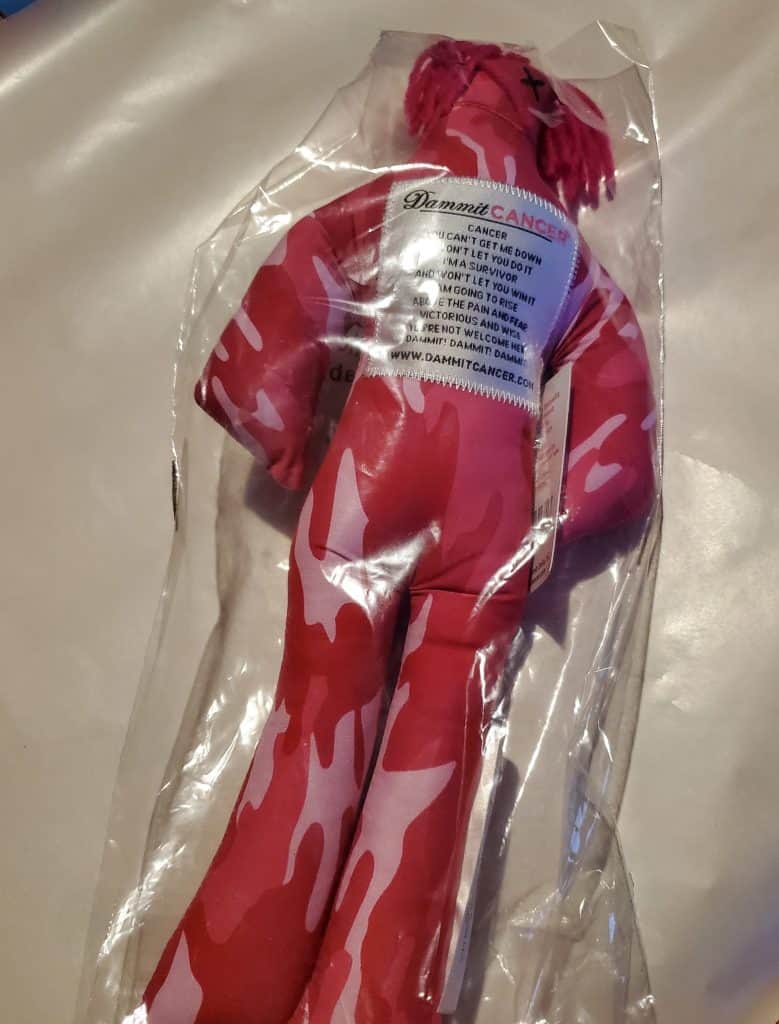 A red and pink camo fabric doll, designed as a thoughtful patient gift, is encased in plastic packaging. Featuring yarn hair and a label with "Dammit Cancer," it offers comforting breast cancer support. The doll sits elegantly on a beige surface, ready to spread warmth and care.