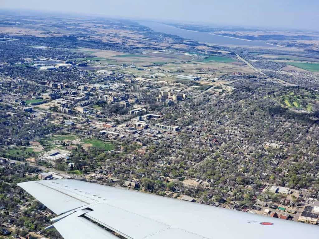 Aerial view of a city with an airplane wing in the foreground, departing from Manhattan KS Airport. The scene includes dense residential areas, commercial buildings, green parks, and surrounding farmlands. A river runs parallel to the horizon, lined by fields and communities. Fly MHK for this picturesque sight!
