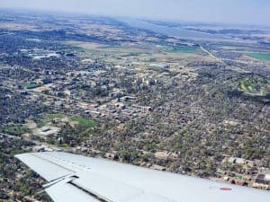 Aerial view of a city with an airplane wing in the foreground, departing from Manhattan KS Airport. The scene includes dense residential areas, commercial buildings, green parks, and surrounding farmlands. A river runs parallel to the horizon, lined by fields and communities. Fly MHK for this picturesque sight!