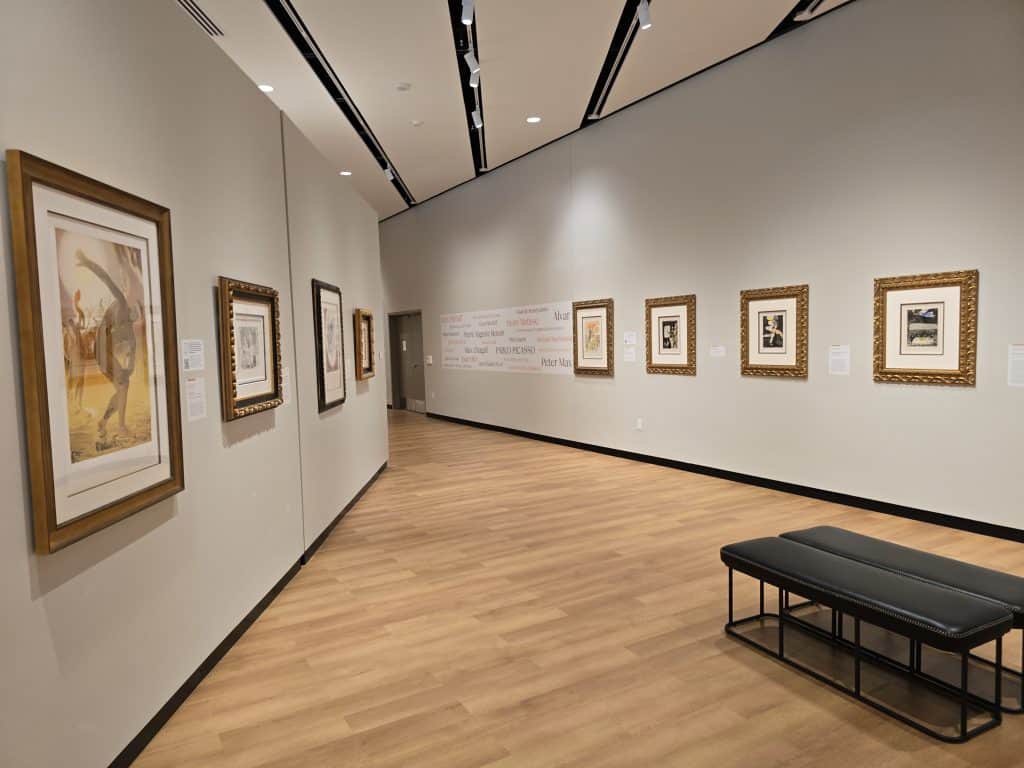 A modern art gallery in the style of the Museum of Art, with light wood floors, white walls, framed paintings and informational plaques lining the space—Art and Light come together with a black cushioned bench inviting visitors to linger.