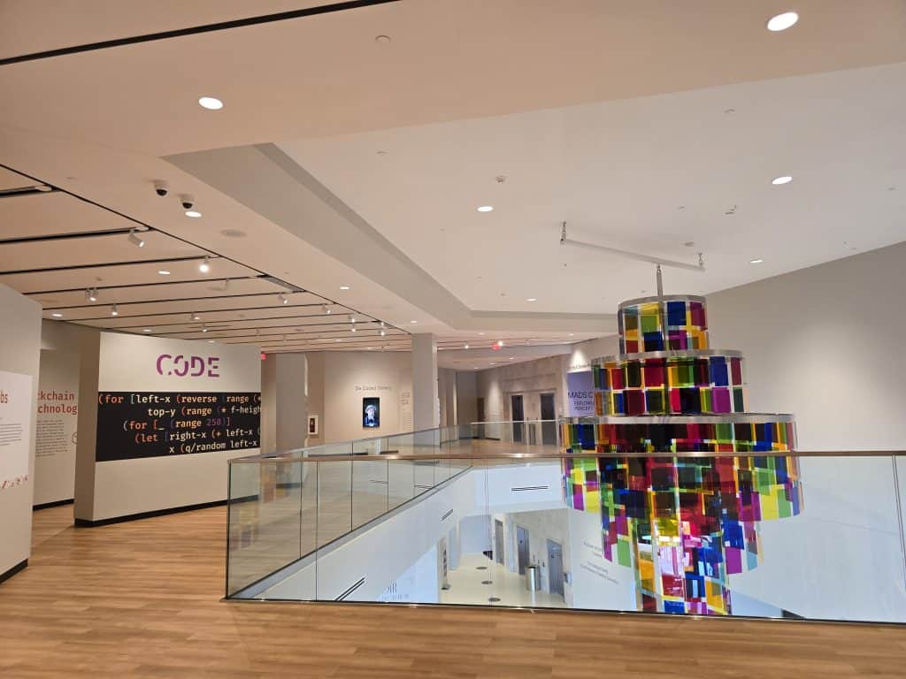 A modern art museum interior in Manhattan KS, featuring light wood floors, white walls with code-themed wall text, and a colorful hanging sculpture of transparent squares above a glass railing at the Museum of Art and Light.