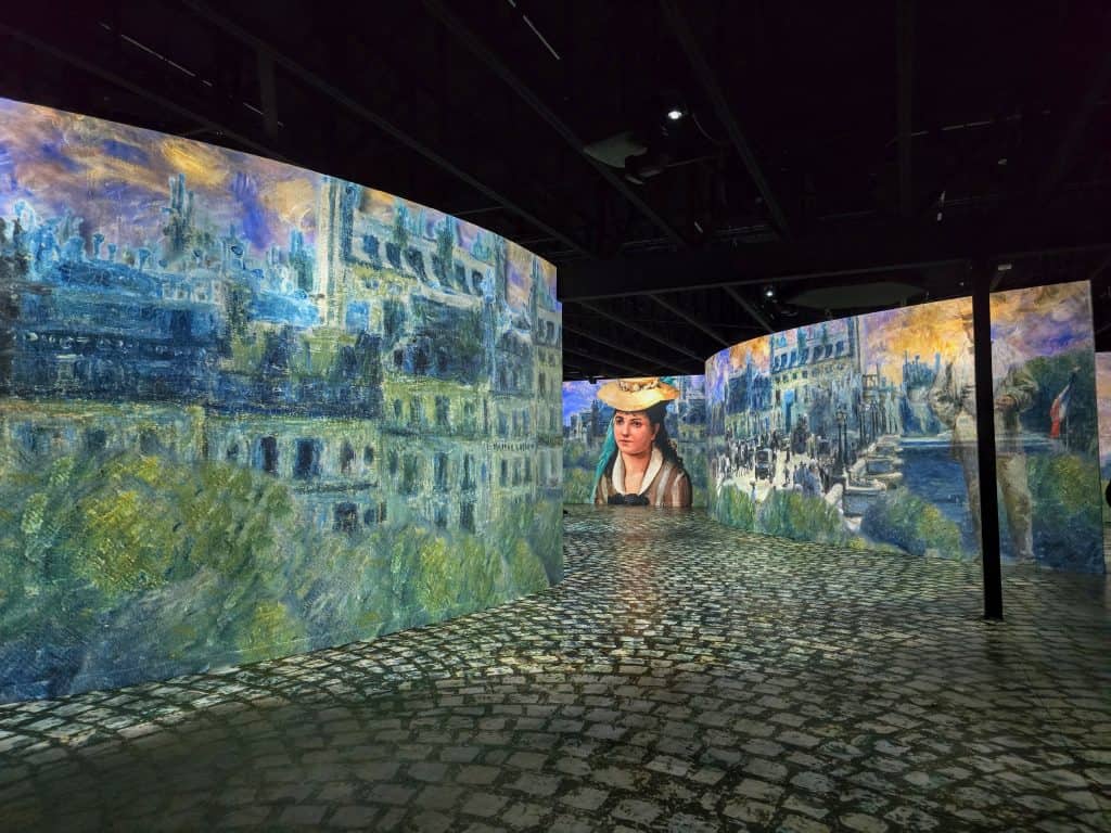 At the Museum of Art and Light in Manhattan KS, large curved screens display impressionist paintings and a portrait of a woman in a hat; the cobblestone-patterned floor creates an immersive art museum exhibit under dim lighting.