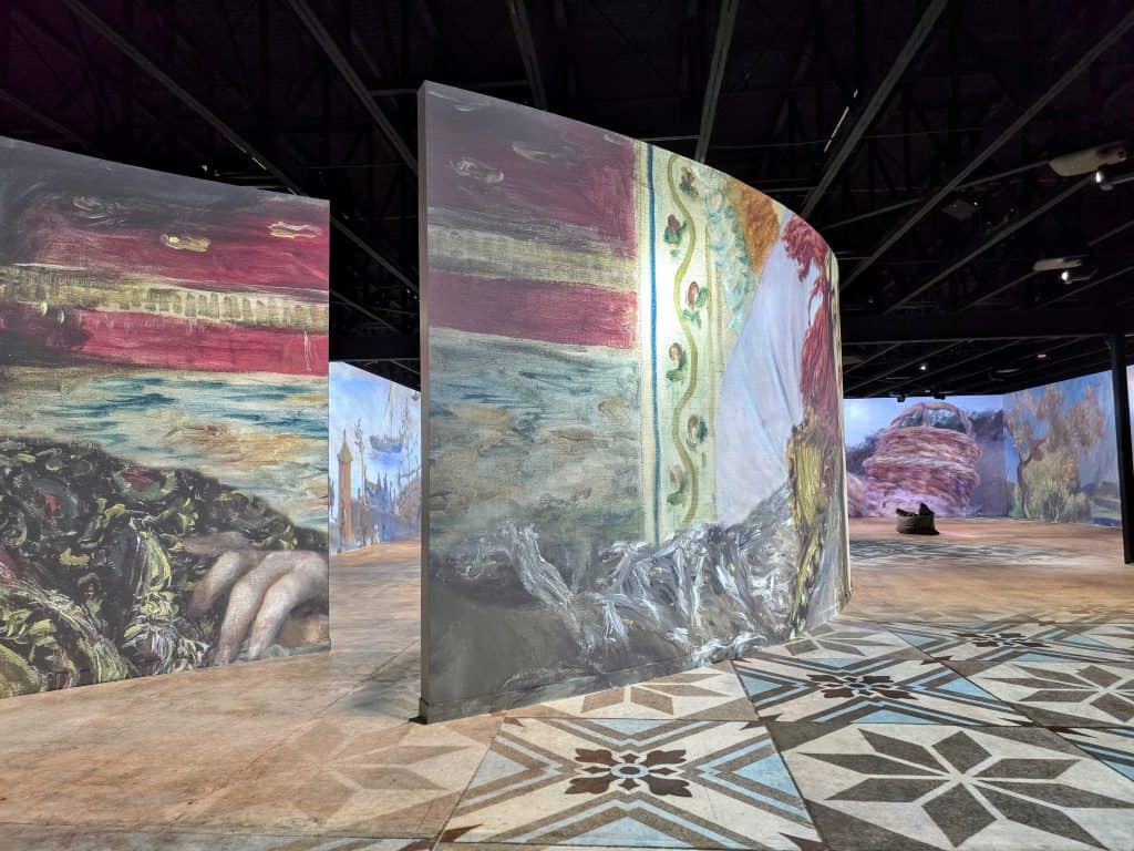Large curved screens display colorful, abstract artworks in the spacious, dimly lit Museum of Art and Light. Patterned floors and scattered visitors create an immersive, interactive exhibition environment in Manhattan KS.