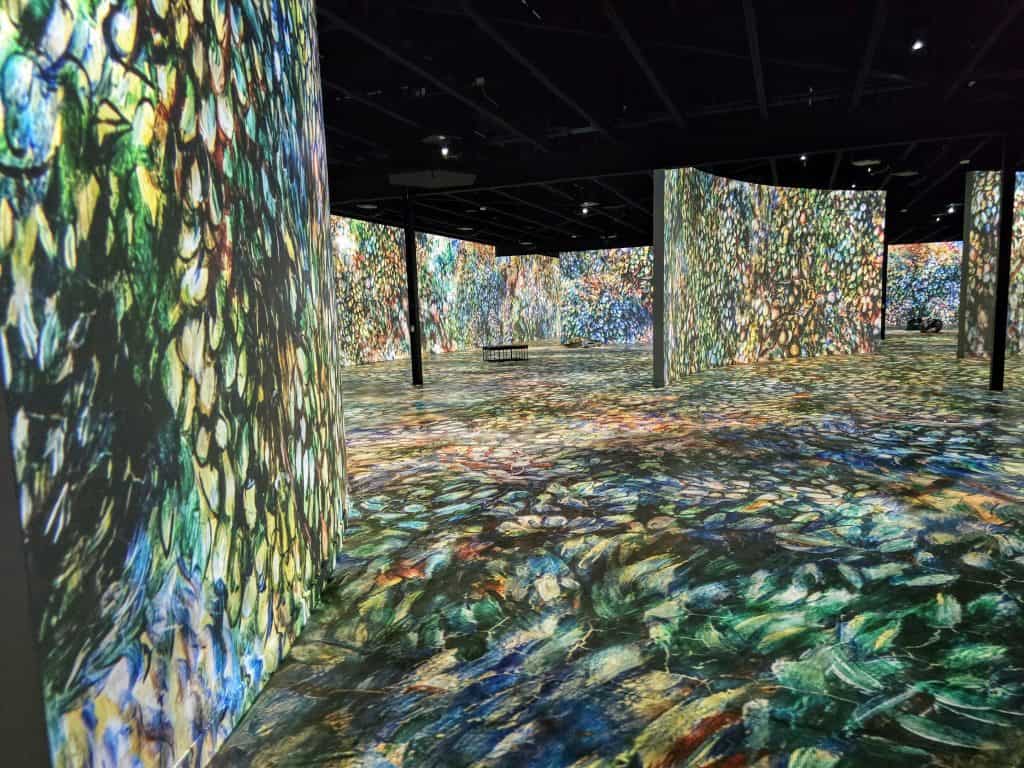 Immersive exhibit room at the Museum of Art and Light in Manhattan KS, featuring large wall and floor projections of colorful, impressionist-style art; dark ceiling with spotlights and a bench visible in the distance.