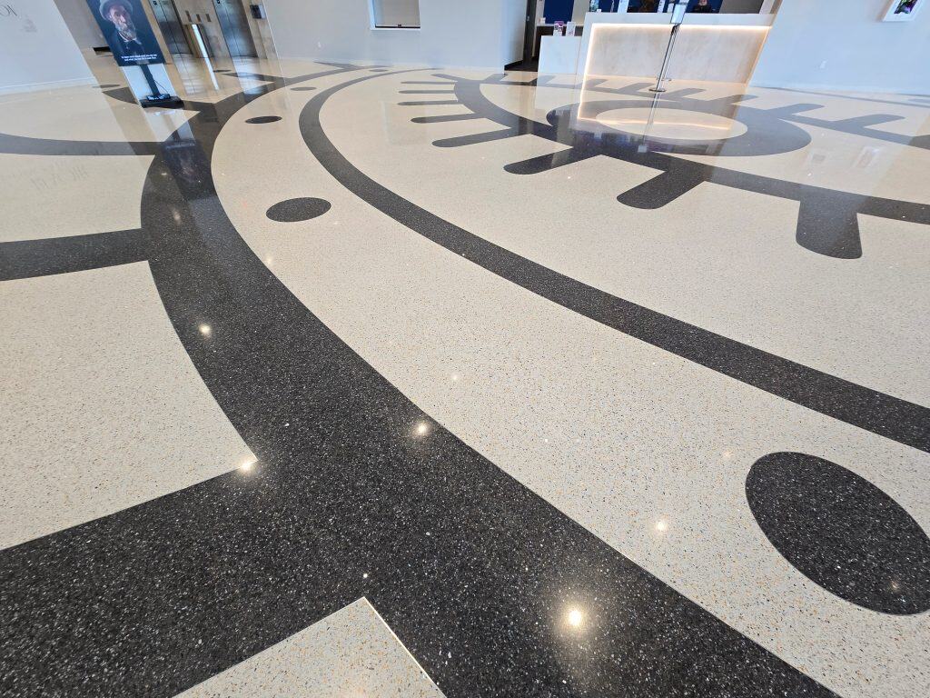 A shiny terrazzo floor features a large, abstract clock design in the modern lobby of the Museum of Art and Light in Manhattan KS, with a reception desk and informational signs visible in the background.