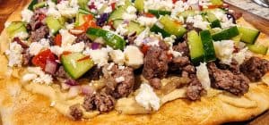 A pizza with meat and vegetables on a wooden cutting board.