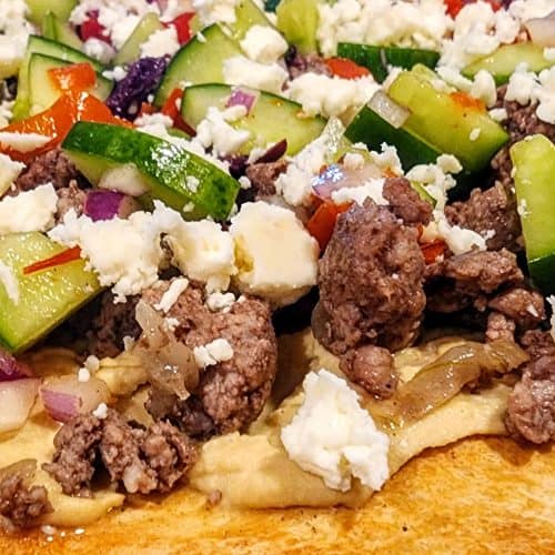 A pizza with meat and vegetables on a wooden cutting board.