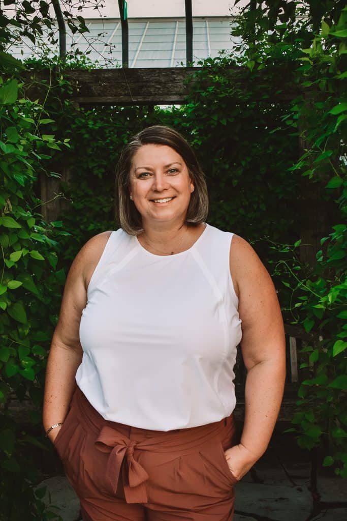 A woman in a white top and brown pants standing in a garden.