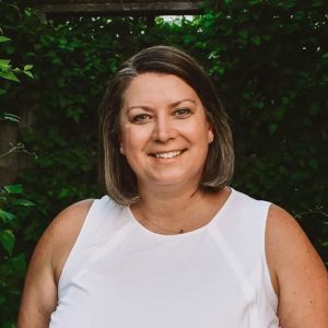 A woman in a white top smiling in front of bushes.