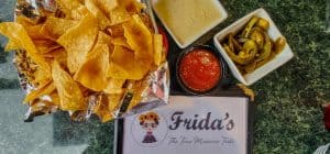 A table with chips, salsa and pickles next to a sign that says frida's.