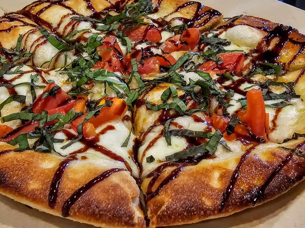 A close-up of a gourmet pizza topped with melted cheese, sliced tomatoes, zucchini, and a drizzle of balsamic glaze, garnished with freshly chopped basil on a thick crust at