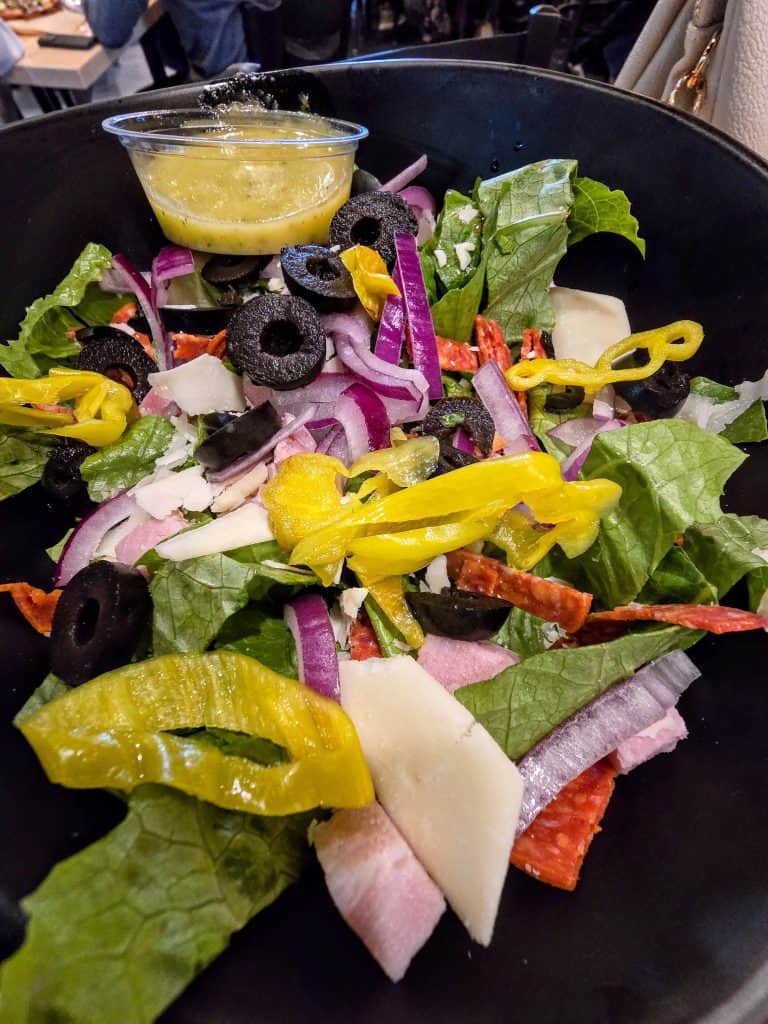 A vibrant garden salad topped with slices of black olives, red onions, strips of yellow bell pepper, cured meats from Highwind Brewing Company, and shaved cheese, served with a side of golden dressing