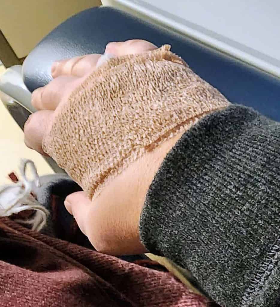 A person's wrist wearing a beige medical wrist brace, resting on a blue armrest, with a reddish-brown blanket draped over their lap.