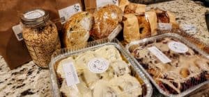 An assortment of baked goods on a countertop, including a jar of local granola, loaves of farm-raised bread wrapped in plastic, and various pastries in clear plastic containers, such as cinnamon rolls and berry-filled pastries. Brown paper bags are also present in the background.