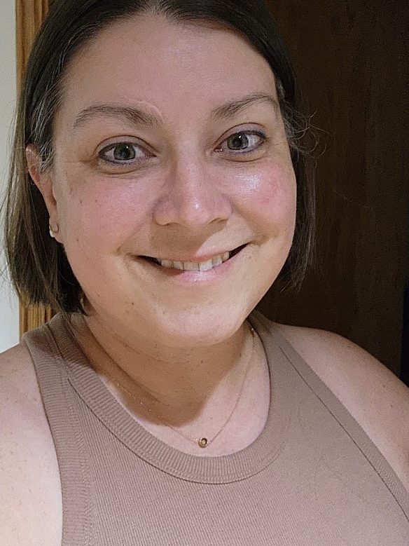 A woman with shoulder-length dark hair and light complexion smiles at the camera, possibly considering her next haircut. She is wearing a sleeveless beige top and a small necklace with a circular pendant, standing in front of a wooden door.