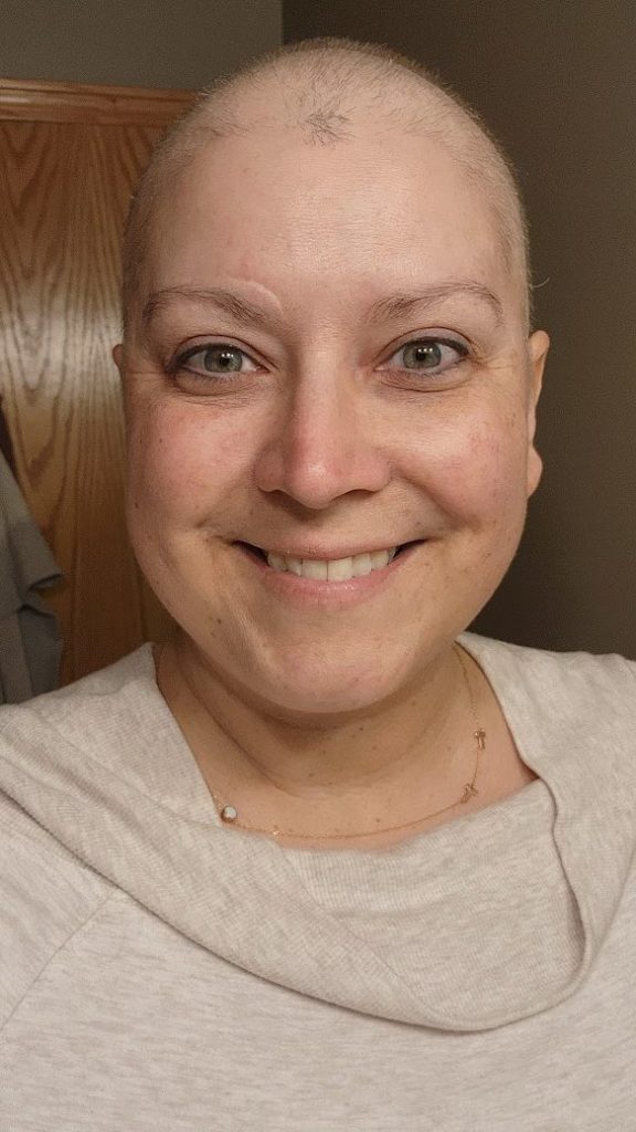 A person with a freshly done haircut smiles warmly at the camera. They are wearing a light-colored top and a delicate necklace. The background shows part of a wooden door and a light-colored wall.