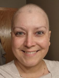 A person with a freshly done haircut smiles warmly at the camera. They are wearing a light-colored top and a delicate necklace. The background shows part of a wooden door and a light-colored wall.