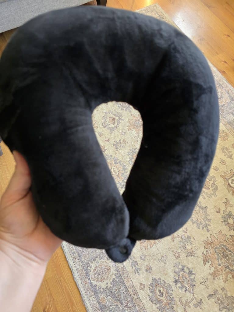 A hand is holding a black U-shaped neck pillow, an essential for making chemo sessions easier. The background features a light-colored rug with intricate patterns and a wooden floor.