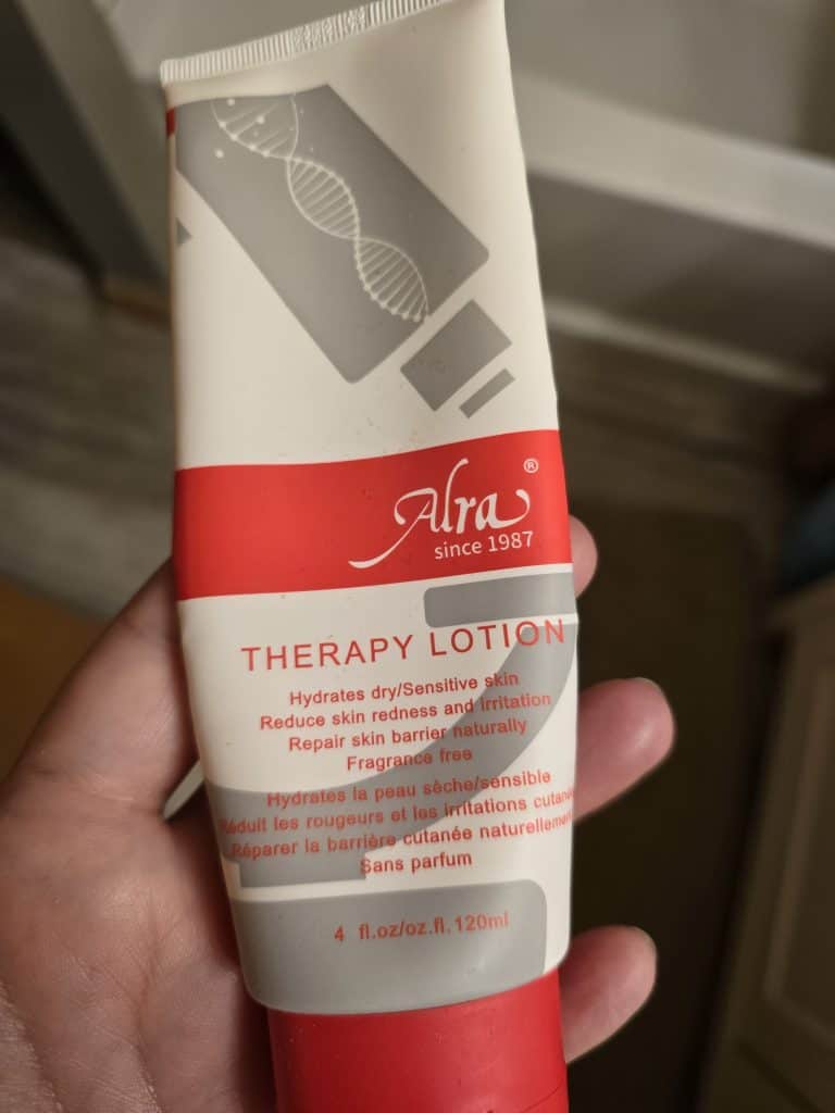 A hand holding a tube of Alra Therapy Lotion. The lotion, in a red and white tube, is designed for dry and sensitive skin, helping to hydrate, reduce redness and irritation, and repair the skin barrier. Especially beneficial for chemo patients, the 4 fl. oz (120 ml) formula makes things easier by soothing troubled skin.
