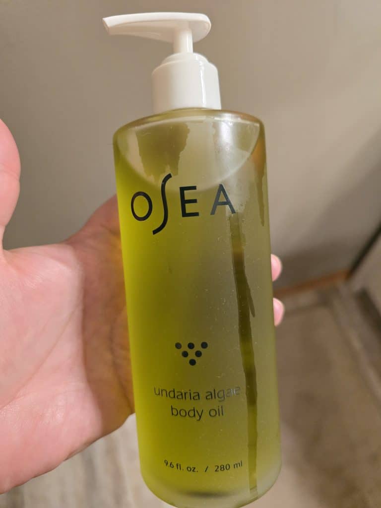 A hand holds a bottle of Osea Undaria Algae Body Oil, perfect for those seeking easier self-care tips. The transparent bottle, filled with green oil and featuring a white pump dispenser, contains 9.6 fl. oz. (280 ml) of product. The background is slightly blurred.