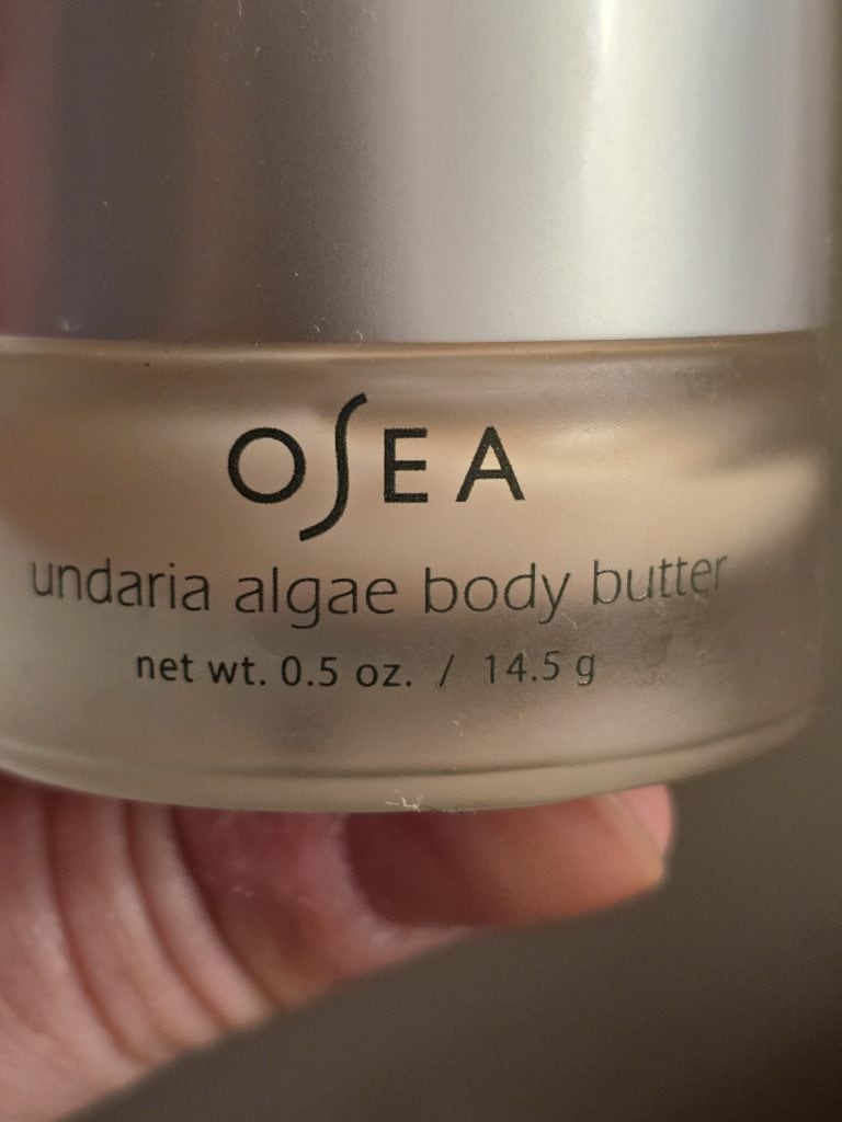 Close-up of a person's finger holding a small jar of Osea Undaria Algae Body Butter, offering a touch of comfort. The jar is clear with a silver lid, displaying the product's name and weight (net wt. 0.5 oz. / 14.5 g) on the label.