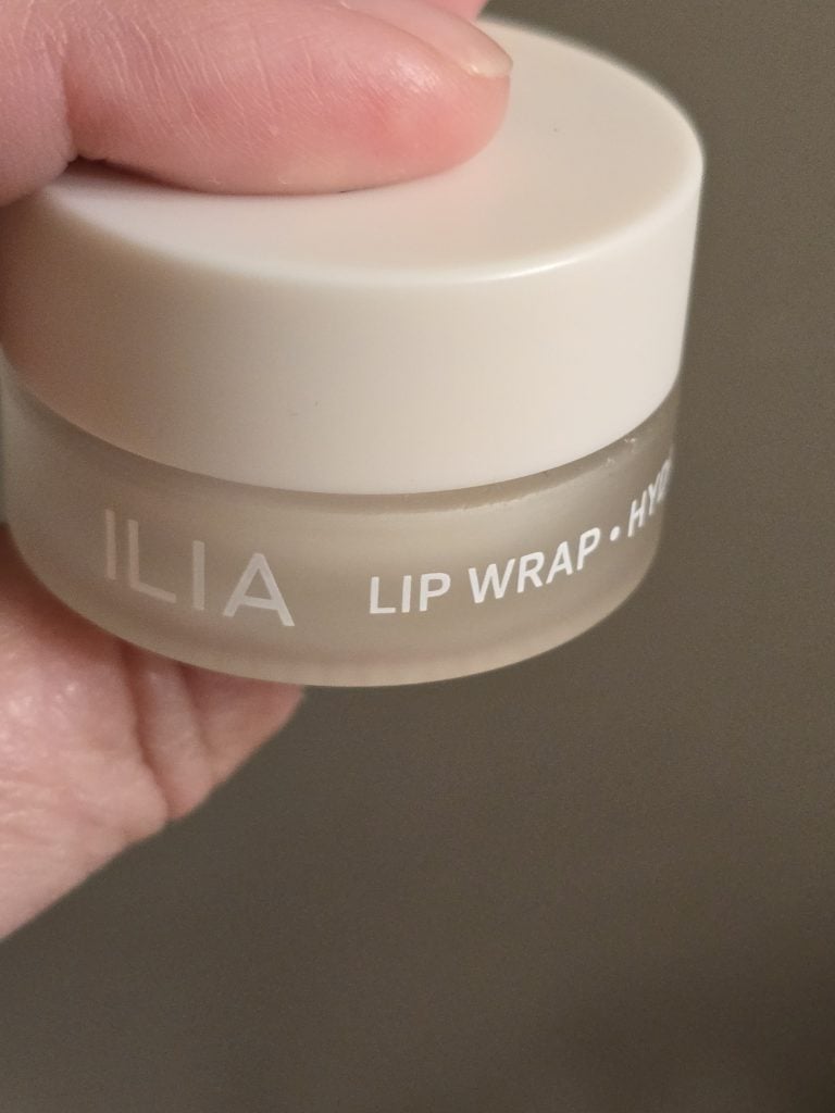 A small, round jar of ILIA Lip Wrap held by a person's hand, offering an easier solution for lip care. The jar has a white lid and the label reads "ILIA LIP WRAP" in white text. The hand is slightly blurred in the background, focusing on the product.