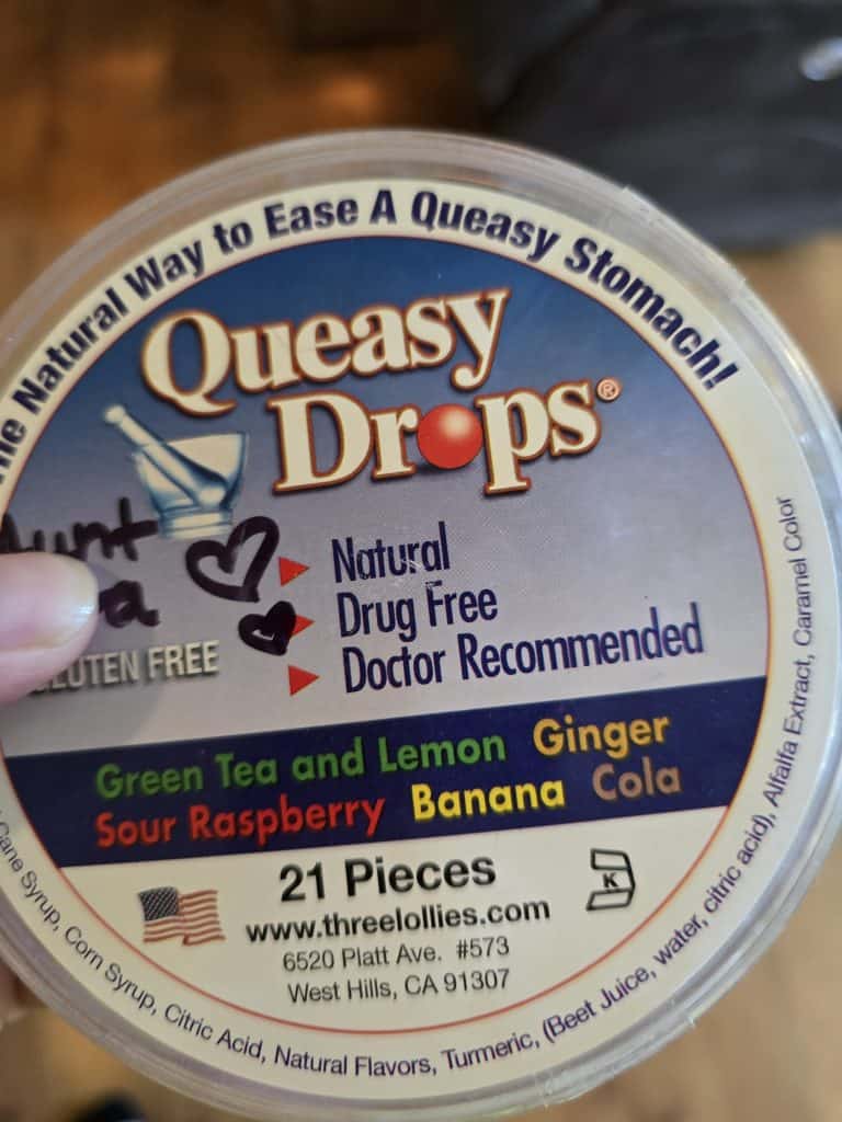 A round container of Queasy Drops is pictured. The label advertises that the product is natural, drug-free, and doctor recommended. These drops can make things easier for those undergoing chemo, with flavors such as Green Tea and Lemon, Ginger, Sour Raspberry, Banana, and Cola.