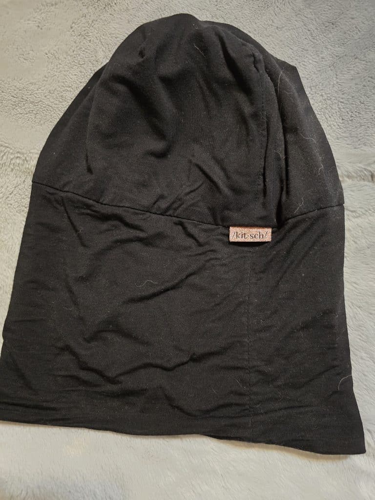 A black slouchy beanie hat lays flat on a soft textured light gray surface. There is a small tag with the brand name "Kitsch" sewn near the bottom edge of the hat. The fabric appears smooth and slightly wrinkled, making it an ideal, comfortable option for those undergoing chemo.