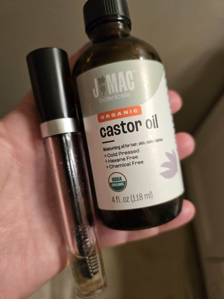 A hand holding a bottle of JMac Botanicals Organic Castor Oil and a mascara tube with a spoolie brush. The castor oil bottle is labeled as moisturizing for hair, skin, nails, and lashes, making post-chemo care easier. It's cold-pressed, hexane-free, and chemical-free.