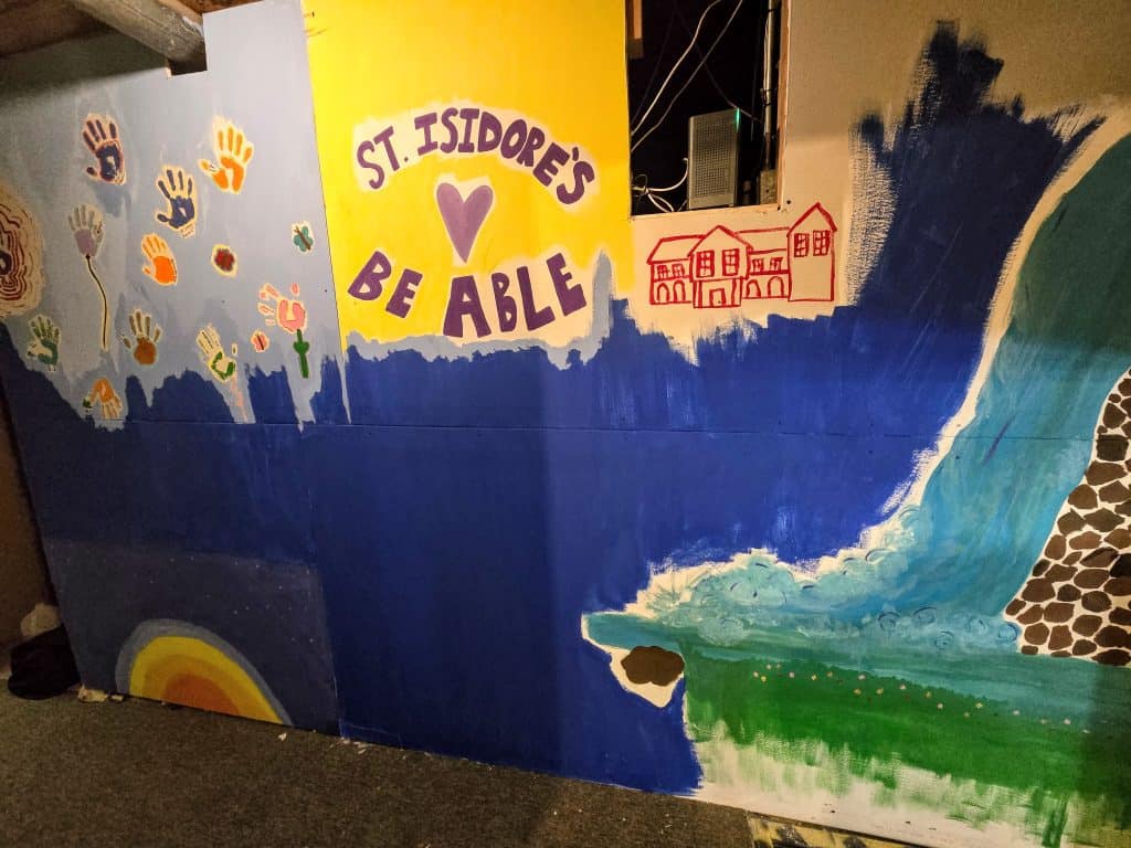 A vibrant mural is displayed in Manhattan, KS, featuring the words "St. Isidore’s BE ABLE" in purple and yellow at the center. Colorful handprints, a rainbow, a house, and various patterns are painted across a blue backdrop, creating an uplifting and creative scene.