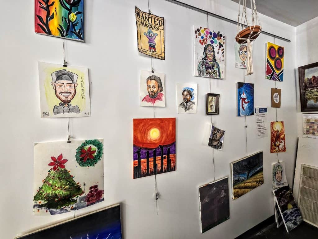 A display of various KS artworks on a white wall, including colorful paintings, portraits, abstract designs, and a Manhattan landscape. The artwork is hung on wires and clips, creating a gallery-like presentation. Some paintings are hung closer to the ceiling and others lower down.