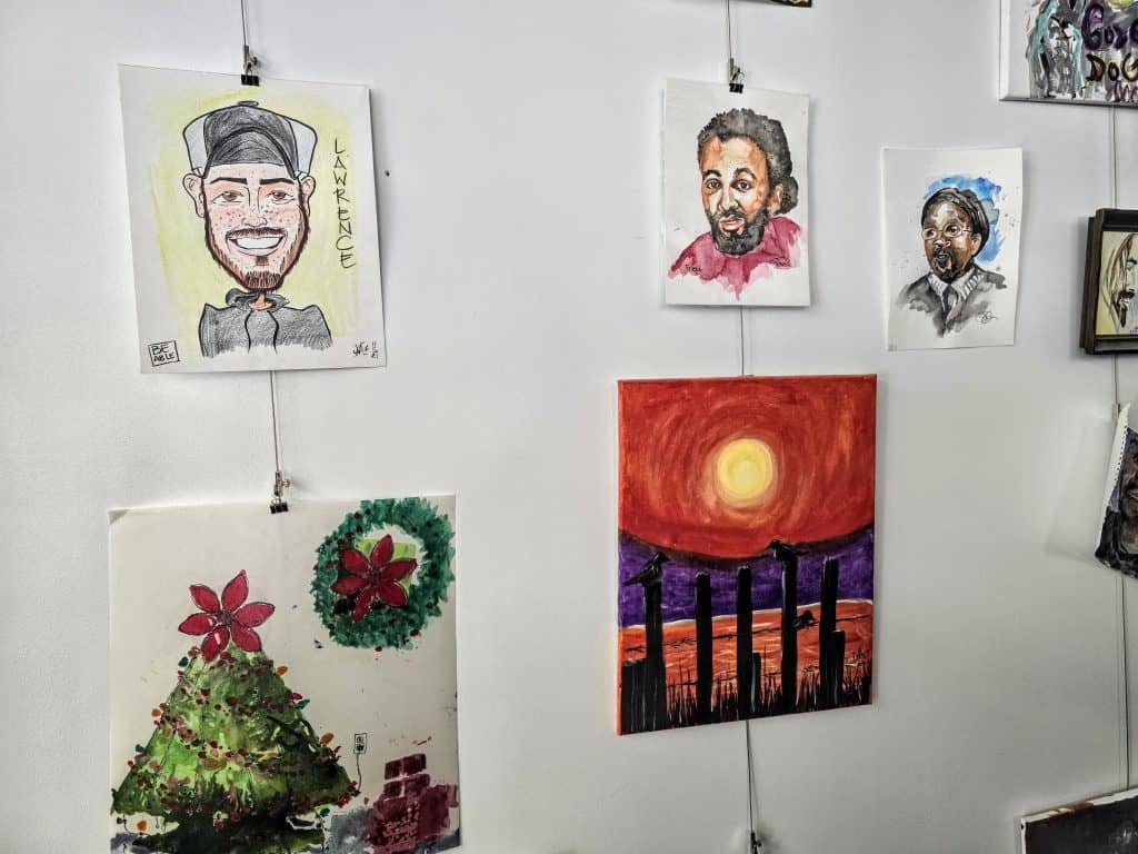 A wall displaying various paintings and drawings. Visible artworks include a caricature of a smiling man in a hat labeled "Lawrence, KS," two portraits of different people, a painting of a sunset with silhouettes of poles, and a festive scene with a decorated Christmas tree.