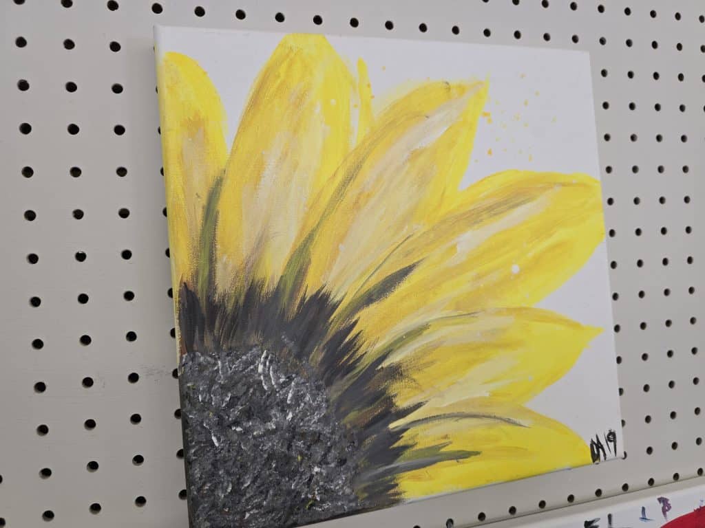 A painting of a sunflower on a white canvas is displayed on a pegboard. The artwork features vibrant yellow petals and a textured black and brown center. The artist's initials, "AH", are signed in the bottom right corner, adding to its charm. This piece would be able to brighten any room in Manhattan, KS.