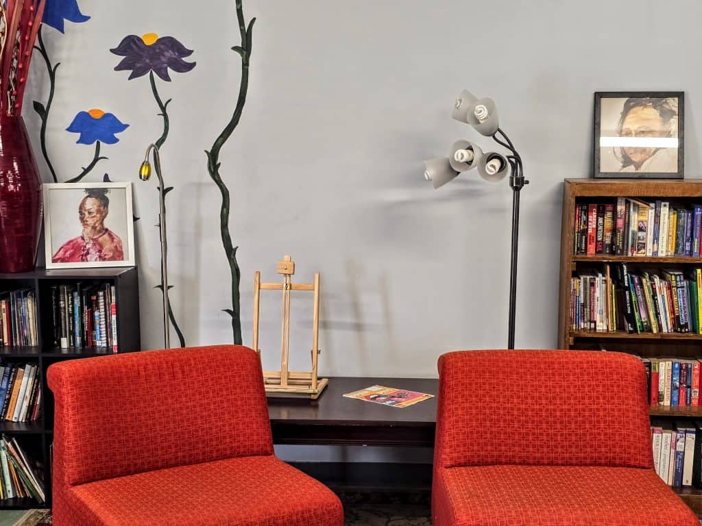 A cozy Manhattan room features two red chairs facing a table holding a magazine. Behind them, a bookcase filled with various books sits to the right. Artwork and a floor lamp decorate the wall, along with a large vase and a wooden easel.