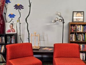A cozy Manhattan room features two red chairs facing a table holding a magazine. Behind them, a bookcase filled with various books sits to the right. Artwork and a floor lamp decorate the wall, along with a large vase and a wooden easel.