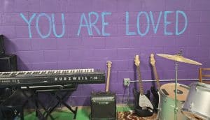 A music room with a keyboard, three guitars, an amplifier, and a drum set, all placed on a purple wall with the message "YOU ARE LOVED" written in large blue letters. In this cozy Manhattan sanctuary, you'll BE ABLE to unleash your musical creativity.