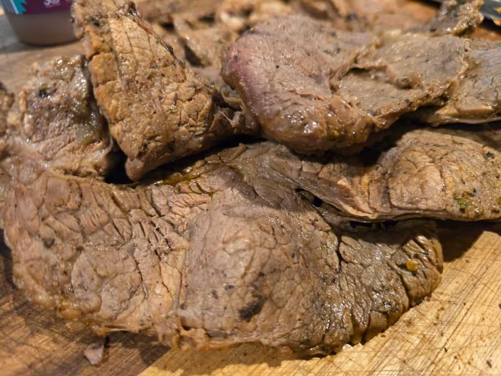 Close-up image of several slices of cooked beef arranged on a wooden cutting board. The meat appears juicy and well-cooked with a slightly charred exterior, showcasing a variety of textures and a rich, brown color—straight from Beto’s Meat Market in Manhattan, KS.
