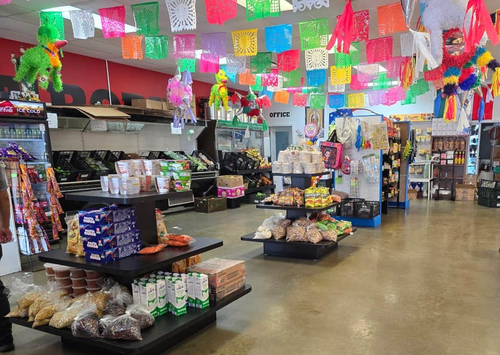 A vibrant store interior at Beto's Meat Market in Manhattan, KS, decorated with colorful papel picado hanging from the ceiling. Shelves display various packaged foods, snacks, and grocery items. A refrigerated section is visible on the left, and piñatas hang near the back of the store.