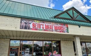 Exterior view of Beto's Meat Market in Manhattan, KS. The banner above the entrance features a picture of a bull and various meats. The storefront has large windows with signs, including one that says "MEAT MARKET" and another that indicates it&rsquo;s open. The building has a green roof.