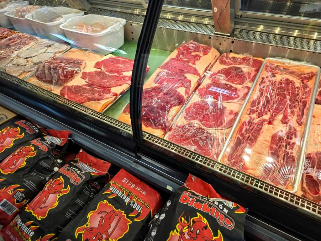 A refrigerated meat display with various cuts of red meat wrapped in plastic at Beto's Meat Market in Manhattan, KS. In front of the display, several bags of "Diablito" charcoal with red and black packaging feature images of a devil.
