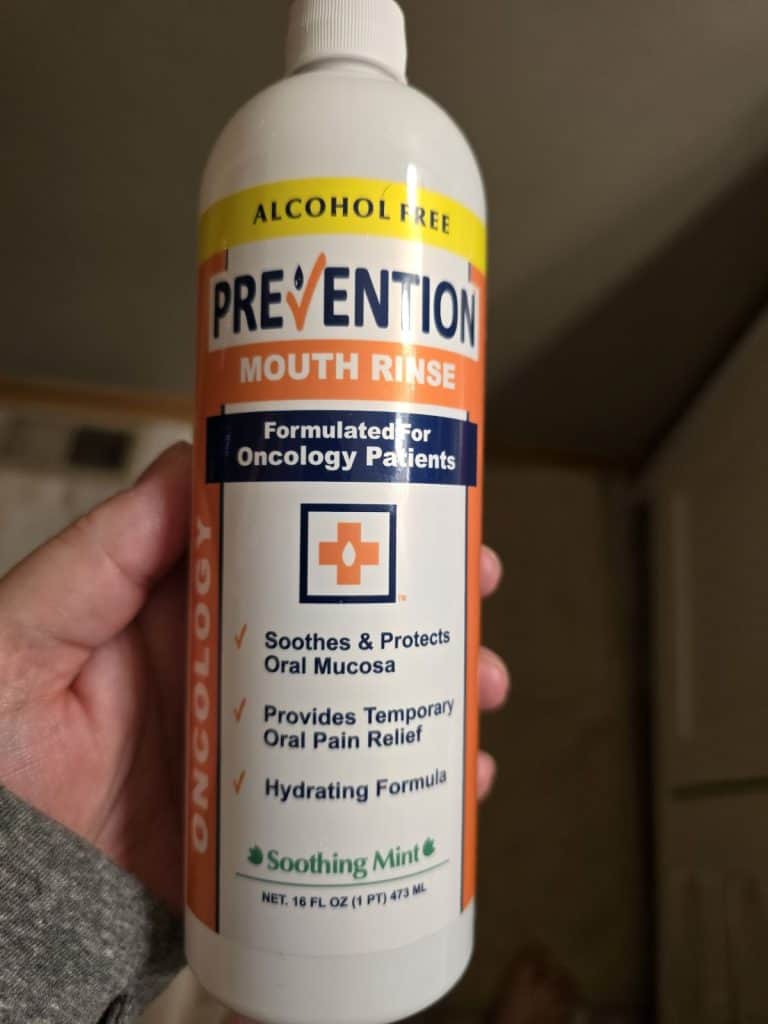 A person is holding a 16 fl oz (473 ml) bottle of alcohol-free Prevention Mouth Rinse, specially formulated for oncology patients undergoing chemo. The label highlights its soothing mint flavor and protective properties for oral mucosa, providing temporary pain relief and hydration—making their routine easier.