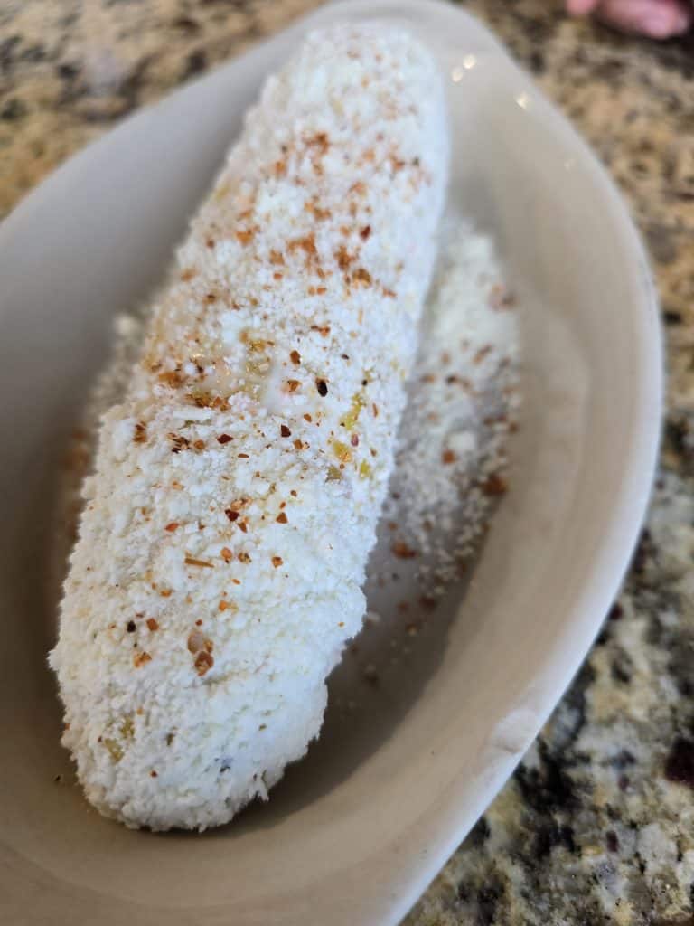 A close-up of a Mexican elote (corn on the cob) topped with mayonnaise, crumbled cheese, and chili powder. Created by Manuel in Hays KS, this fusion dish is served on a white, oval-shaped dish placed on a granite countertop.