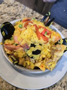 A bowl of colorful paella sits on a white plate. The dish is filled with yellow saffron rice, shrimp, mussels, red bell pepper strips, peas, and chunks of chicken and pork. This fusion of flavors is presented on a speckled granite countertop in Manuel Hays' kitchen.