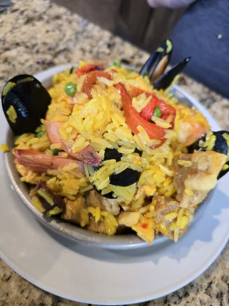 A bowl of colorful paella sits on a white plate. The dish is filled with yellow saffron rice, shrimp, mussels, red bell pepper strips, peas, and chunks of chicken and pork. This fusion of flavors is presented on a speckled granite countertop in Manuel Hays' kitchen.