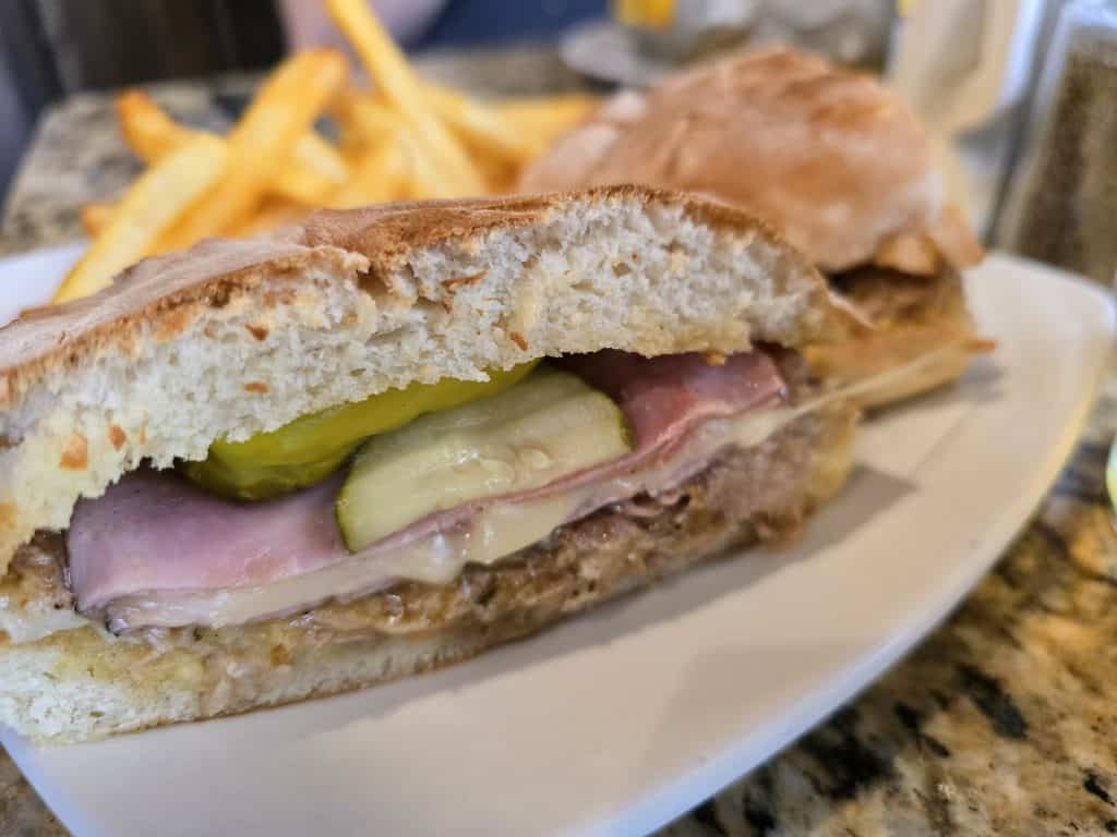 Close-up of a Fusion by Manuel sandwich in Hays, KS, featuring slices of ham, pickles, cheese, and some type of meat on a crusty bread roll. Served on a white plate with French fries in the background.