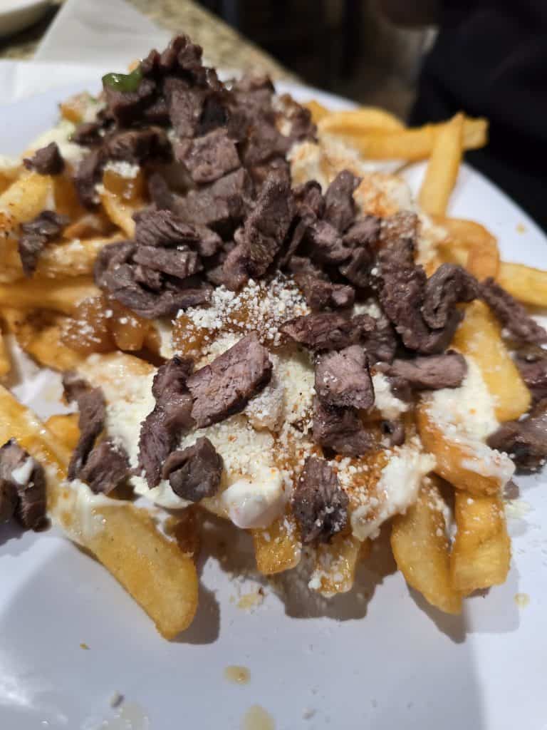 A close-up of a plate of loaded fries topped with grilled steak, melted cheese, and a sprinkle of grated cheese. The fries are golden and crispy, with generous toppings. This fusion dish by Manuel in Hays, KS appears appetizing and hearty.